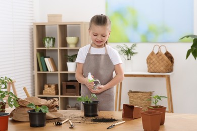 Cute little girl spraying seedling in pot at wooden table in room