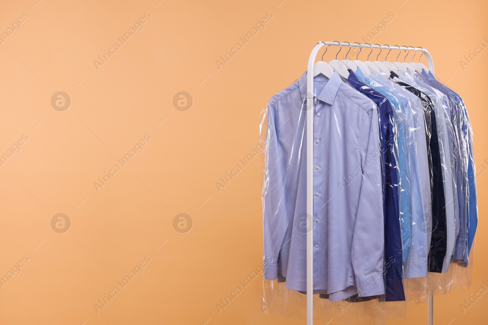 Photo of Dry-cleaning service. Many different clothes in plastic bags hanging on rack against beige background, space for text