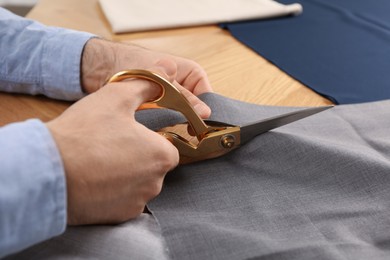 Photo of Man cutting grey fabric with scissors at wooden table, closeup