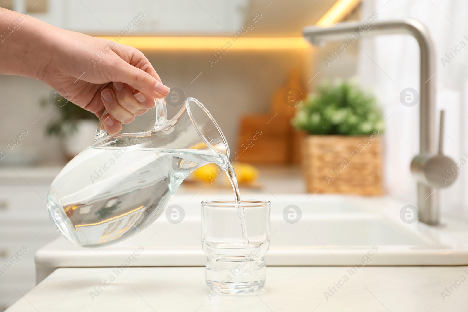 Photo of Woman pouring water from jug into glass at white table in kitchen, closeup. Space for text