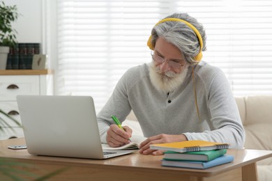 Photo of Middle aged man with laptop, notebook and headphones learning at table indoors