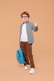 Photo of Happy schoolboy with backpack showing thumb up on beige background