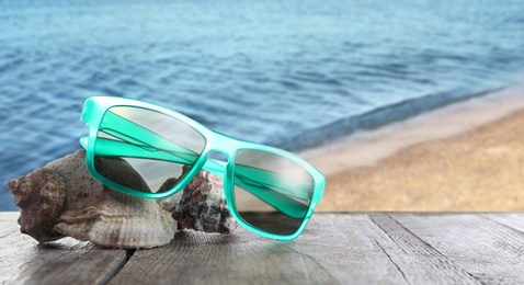 Shells and stylish sunglasses on wooden table near sea with sandy beach