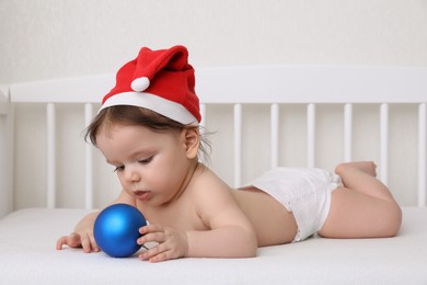 Photo of Cute little baby wearing Santa hat with Christmas ball in crib at home