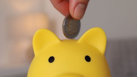 Photo of Woman putting coin into yellow piggy bank on blurred background, closeup