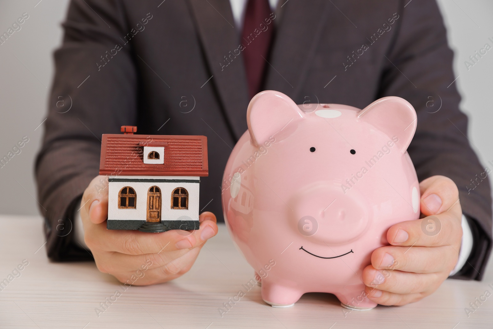 Photo of Man holding house model and piggy bank at wooden table, closeup. Saving money concept