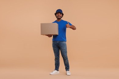 Photo of Happy young courier with parcel showing thumb up on light brown background
