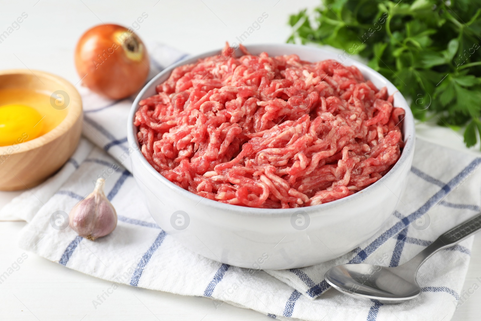 Photo of Raw ground meat in bowl, garlic, egg, parsley and spoon on white table, closeup