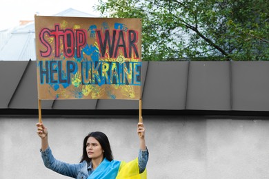 Photo of Sad woman wrapped in Ukrainian flag holding placard with words Stop War Help Ukraine outdoors. Space for text