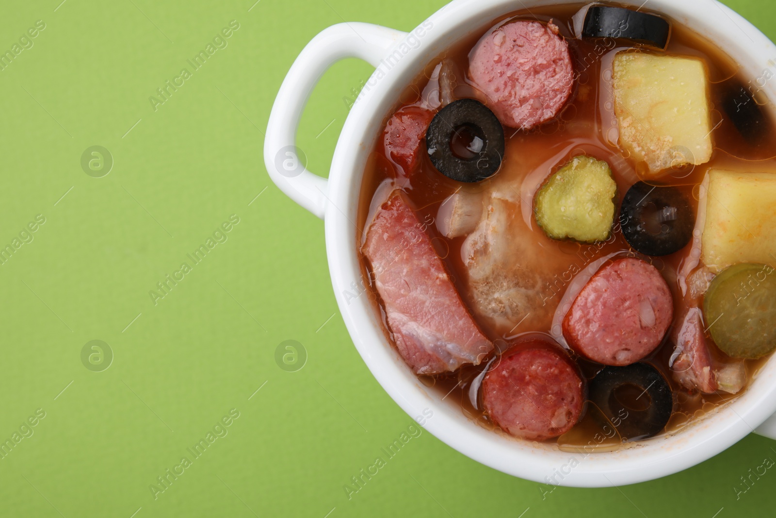 Photo of Meat solyanka soup with thin dry smoked sausages in pot on light green background, top view. Space for text