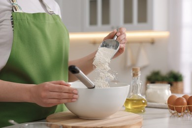 Photo of Making bread. Woman putting flour into bowl at white table in kitchen, closeup