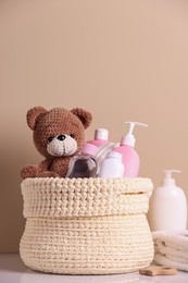 Photo of Knitted basket with baby cosmetic products and toy bear on white table against beige background