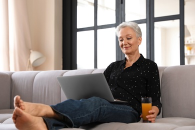 Photo of Mature woman with laptop and drink sitting on sofa at home. Smart aging