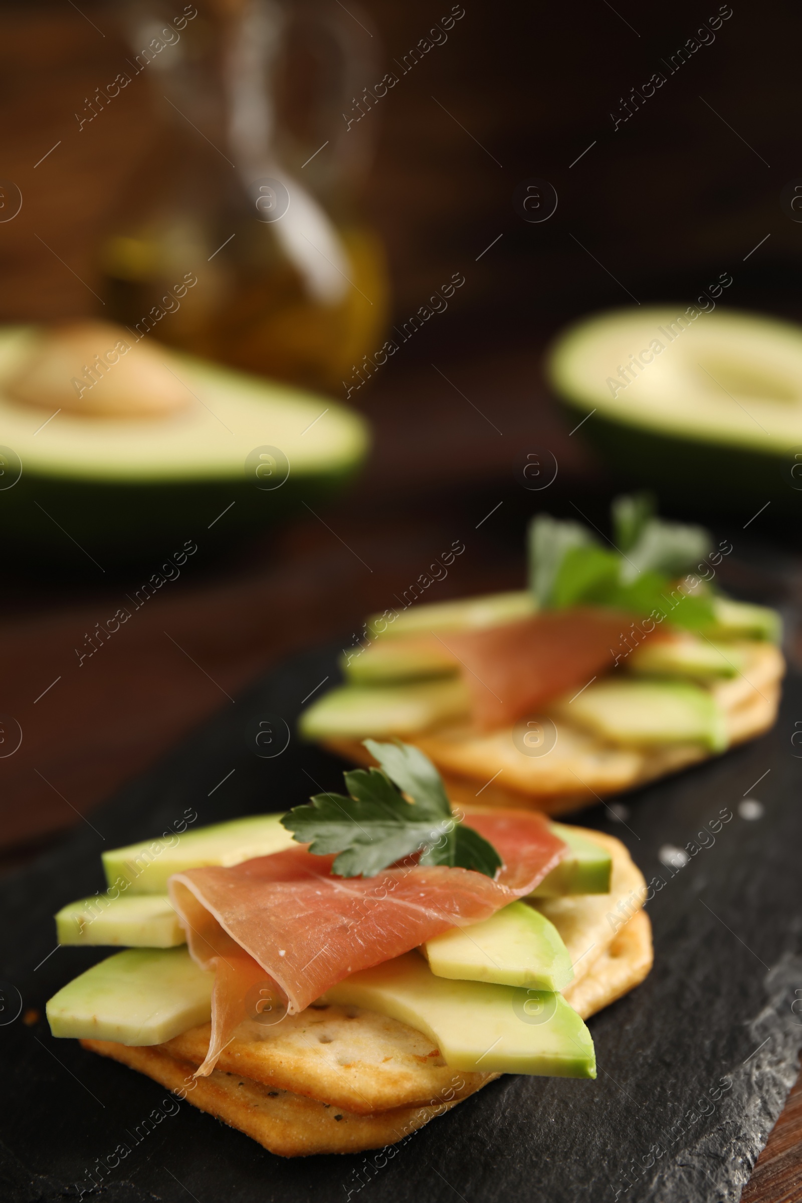 Photo of Delicious crackers with avocado, prosciutto and parsley on slate board, closeup