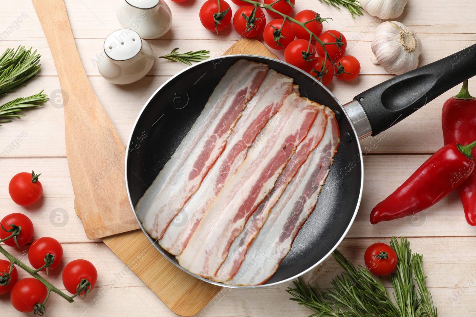 Photo of Slices of raw bacon in frying pan and fresh products on wooden table, flat lay