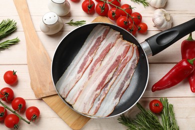 Photo of Slices of raw bacon in frying pan and fresh products on wooden table, flat lay