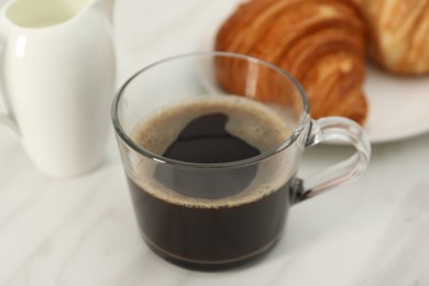 Photo of Aromatic coffee in glass cup, pitcher and fresh croissants on white marble table, closeup