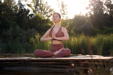 Photo of Beautiful woman practicing Padmasana on yoga mat on wooden pier near pond. Lotus pose