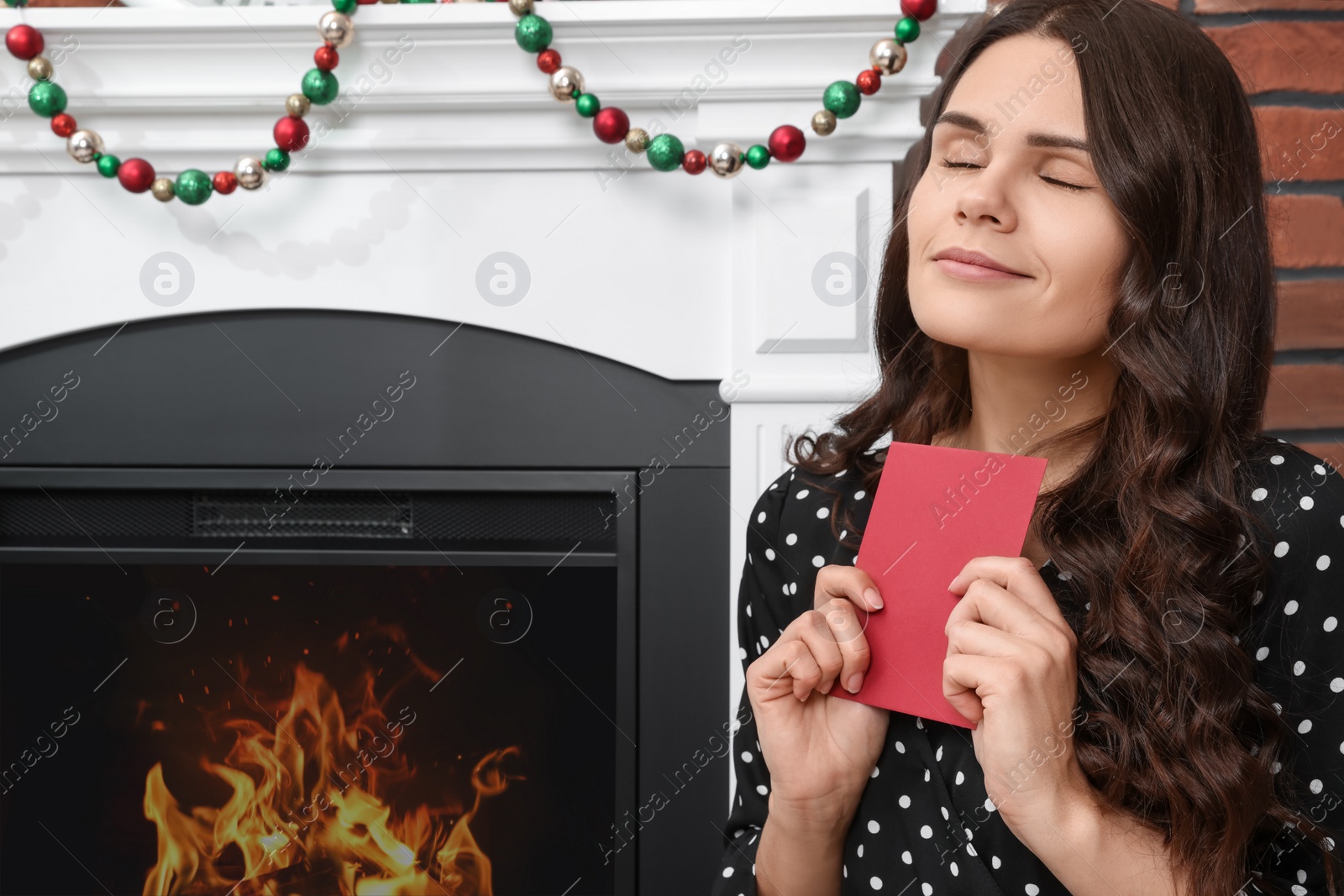 Photo of Young woman with greeting card sitting near fireplace indoors