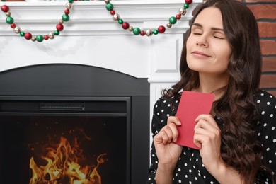Young woman with greeting card sitting near fireplace indoors