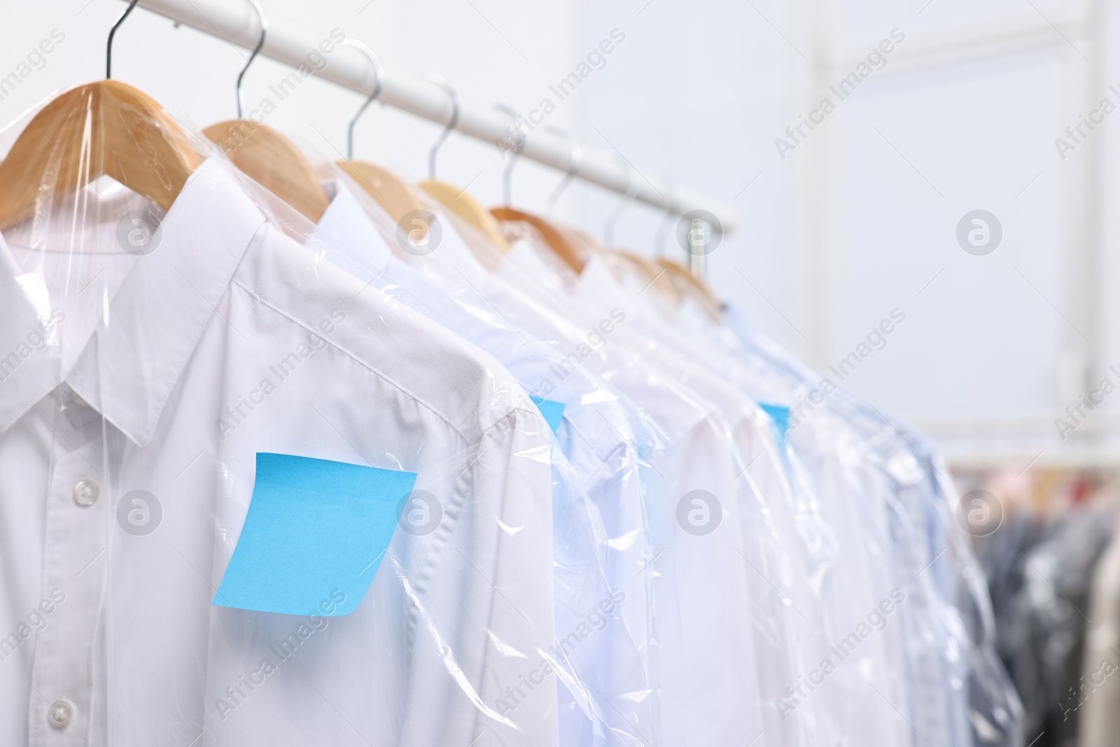 Photo of Dry-cleaning service. Many different clothes in plastic bags hanging on rack indoors, closeup