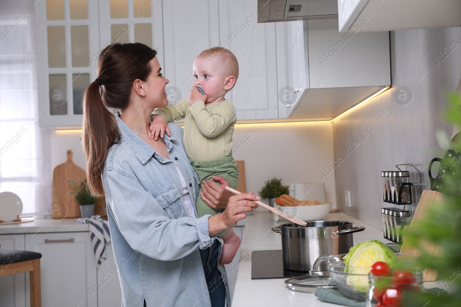 Photo of Happy young woman holding her cute little baby while cooking in kitchen