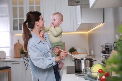 Happy young woman holding her cute little baby while cooking in kitchen