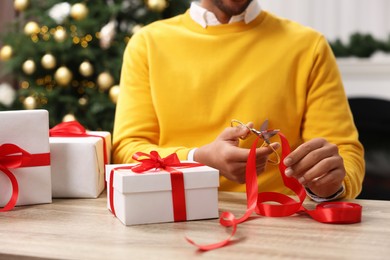 Christmas gift. Man cutting red ribbon at wooden table indoors, closeup