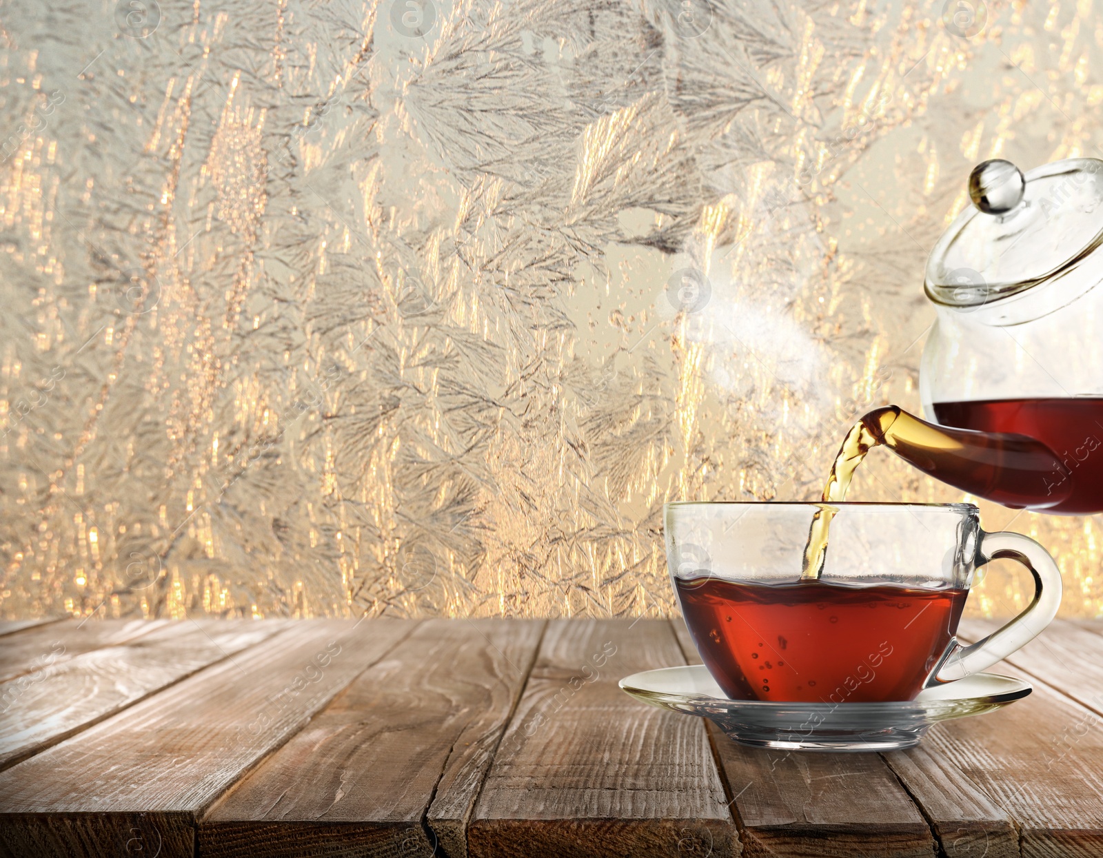 Image of Pouring tea into glass cup on wooden table near window covered with frost 