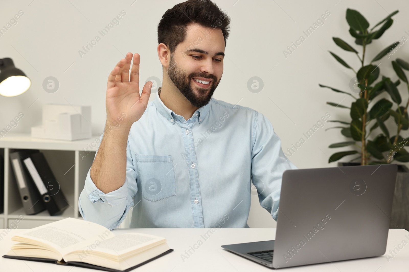 Photo of Young man waving hello during video chat via laptop at table indoors