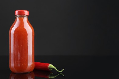 Photo of Spicy chili sauce in bottle and pepper against dark background, space for text