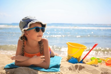 Little girl in stylish sunglasses and hat sunbathing on sandy beach near sea, space for text