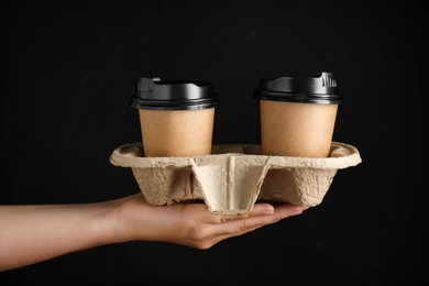 Woman holding cardboard holder with takeaway paper coffee cups on black background, closeup