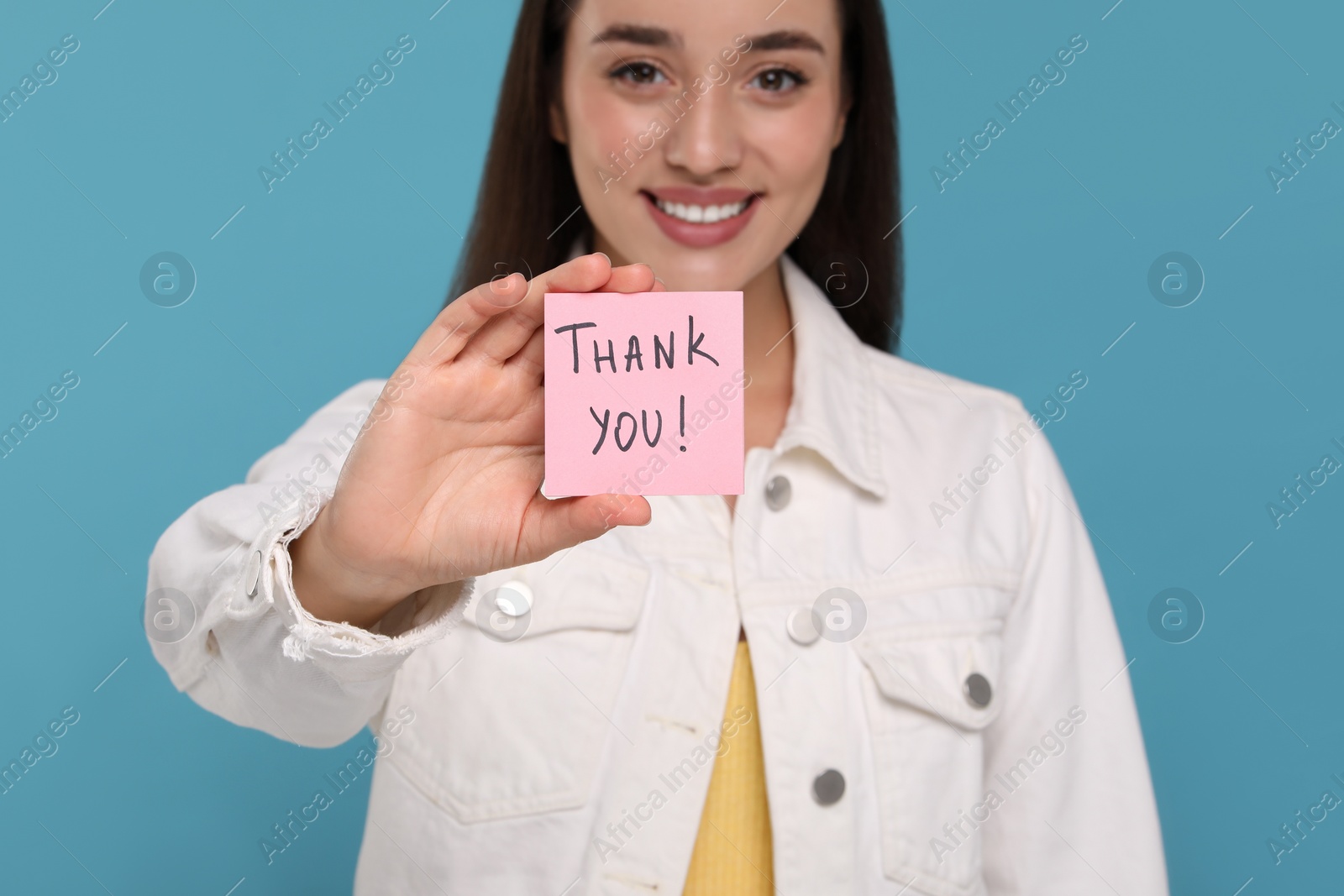 Photo of Happy woman holding paper note with phrase Thank You on light blue background, selective focus