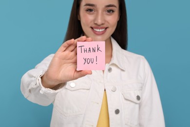 Photo of Happy woman holding paper note with phrase Thank You on light blue background, selective focus
