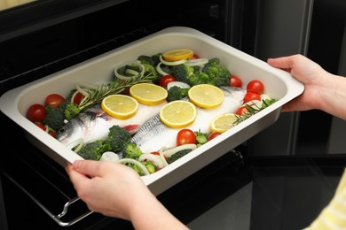 Photo of Woman putting baking dish with raw fish and vegetables into oven, closeup