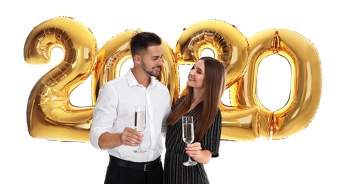 Photo of Happy young couple with glasses of champagne near golden 2020 balloons on white background. New Year celebration