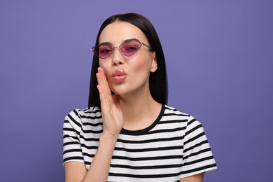 Photo of Beautiful young woman in stylish sunglasses blowing kiss on purple background