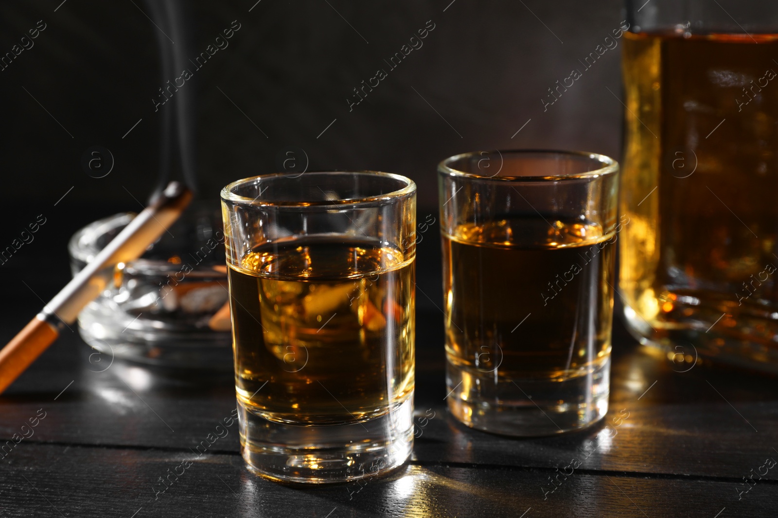 Photo of Alcohol addiction. Whiskey in glasses, smoldering cigarette and ashtray on black wooden table, closeup