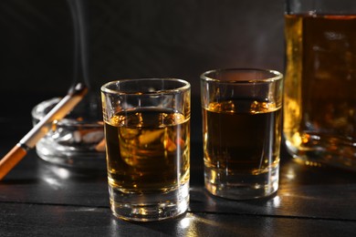 Photo of Alcohol addiction. Whiskey in glasses, smoldering cigarette and ashtray on black wooden table, closeup