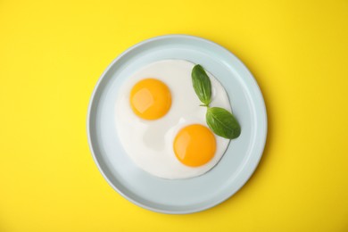 Photo of Tasty fried eggs with basil in plate on yellow background, top view