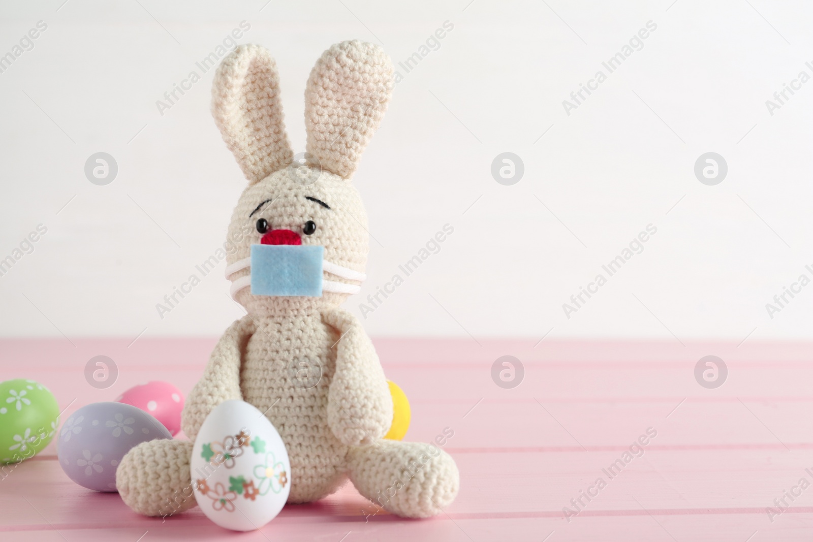 Photo of Toy bunny with protective mask and painted eggs on pink wooden table, space for text. Easter holiday during COVID-19 quarantine