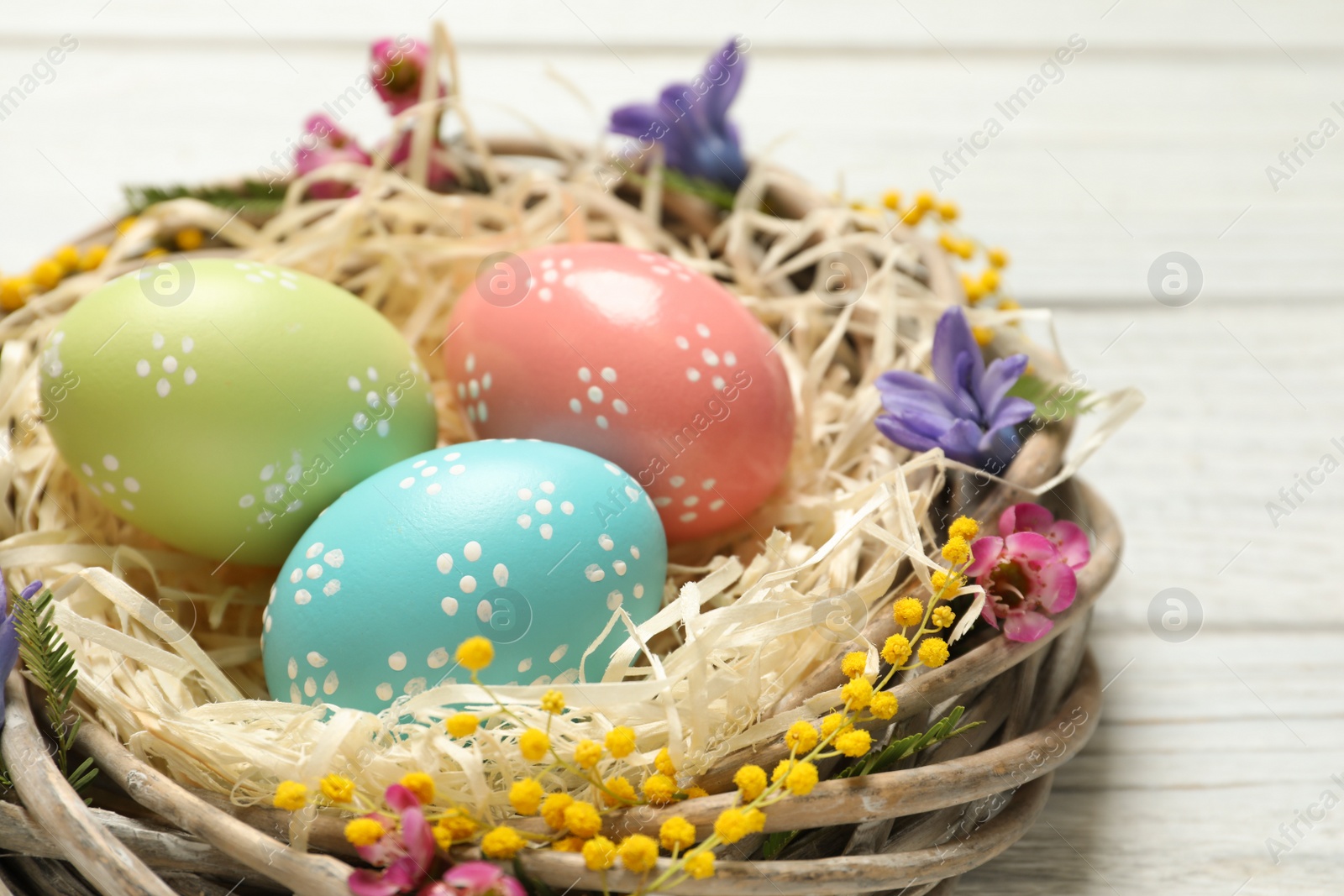 Photo of Wicker nest with painted Easter eggs and flowers on table, closeup