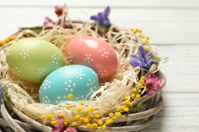 Wicker nest with painted Easter eggs and flowers on table, closeup