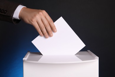 Man putting his vote into ballot box on dark blue background, closeup