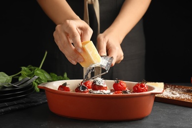 Woman grating cheese onto baked eggplant with tomatoes at black table, closeup