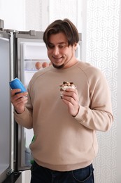 Photo of Overweight man holding dessert and tin can with beverage near open refrigerator in kitchen