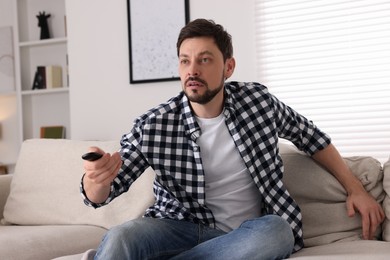 Photo of Man changing TV channels with remote controller on sofa at home