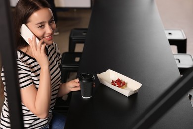 Happy young woman with paper cup of coffee talking on smartphone at table in hostel dining room