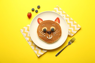 Photo of Creative serving for kids. Plate with cute cat made of pancakes, berries, cream, banana and chocolate paste on yellow background, flat lay